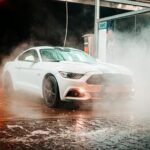 White Ford Mustang in a misty carwash setting, illuminated at night in an urban environment.