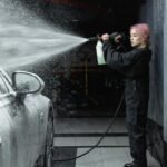 Young woman cleaning a car with a high-pressure hose inside a garage, creating a splash of water and foam.