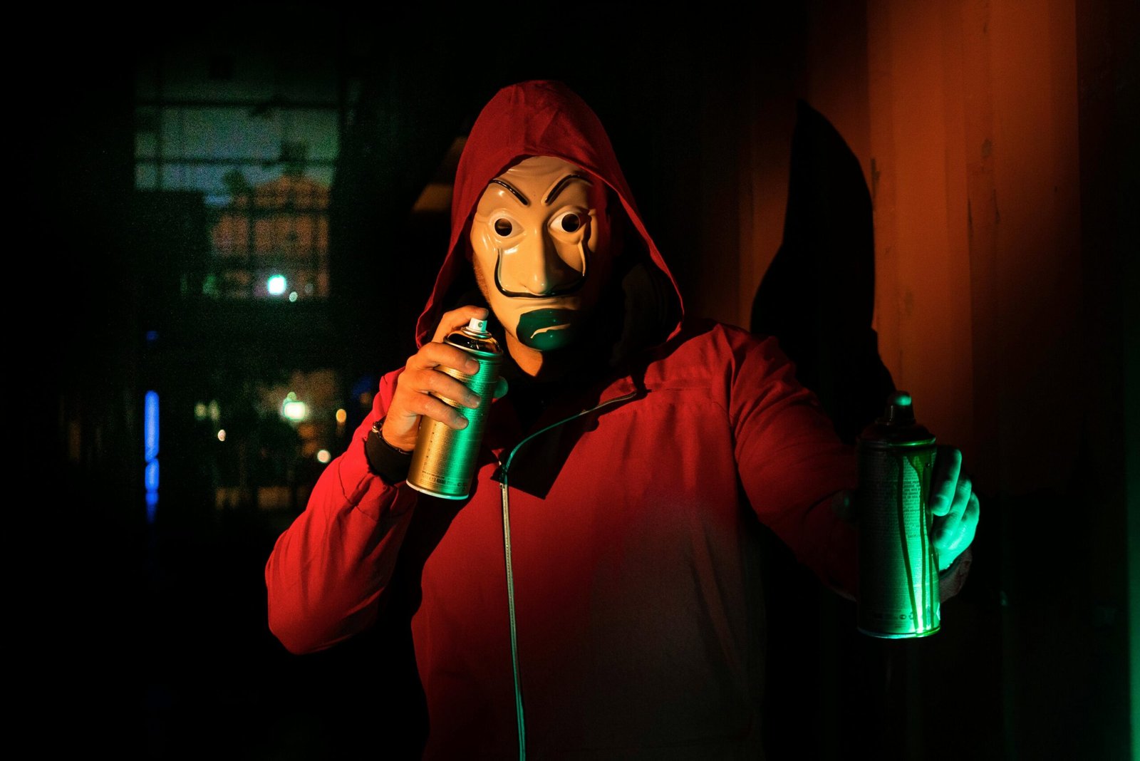 A masked person in a red hoodie holds spray paints, creating street art at night.